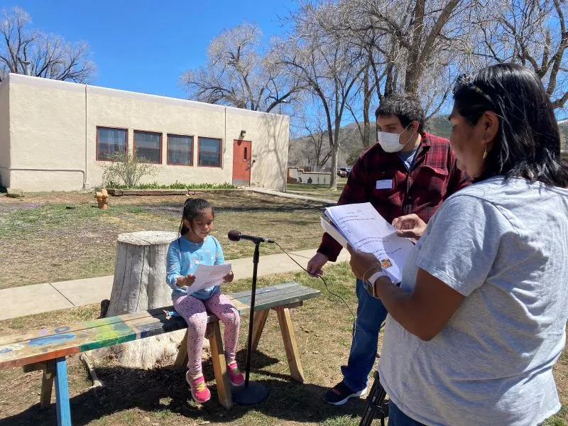 Girl Student at Taos Day School Outside Sitting on a Bench in Front of a Microphone Reading a Poem for the "I Am From Poem" Project with a Man and a Woman Offering Support.