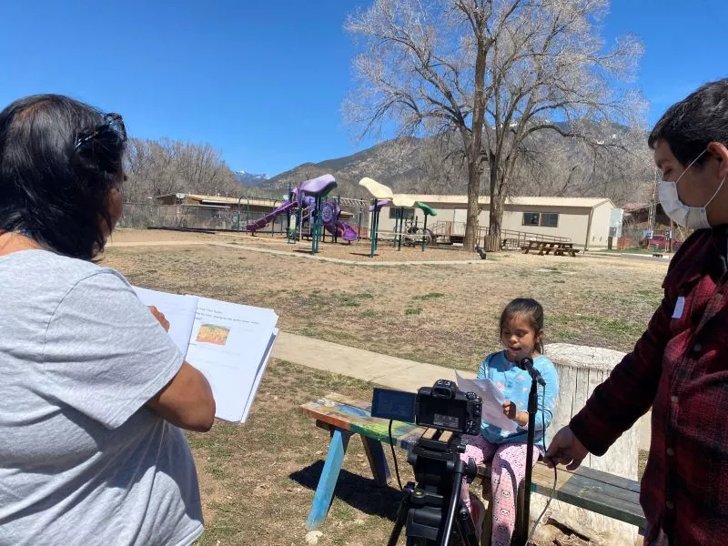 Girl Student at Taos Day School Outside Sitting on a Bench in Front of a Microphone Reading a Poem for the "I Am From Poem" Project with a Man and a Woman Offering Support.