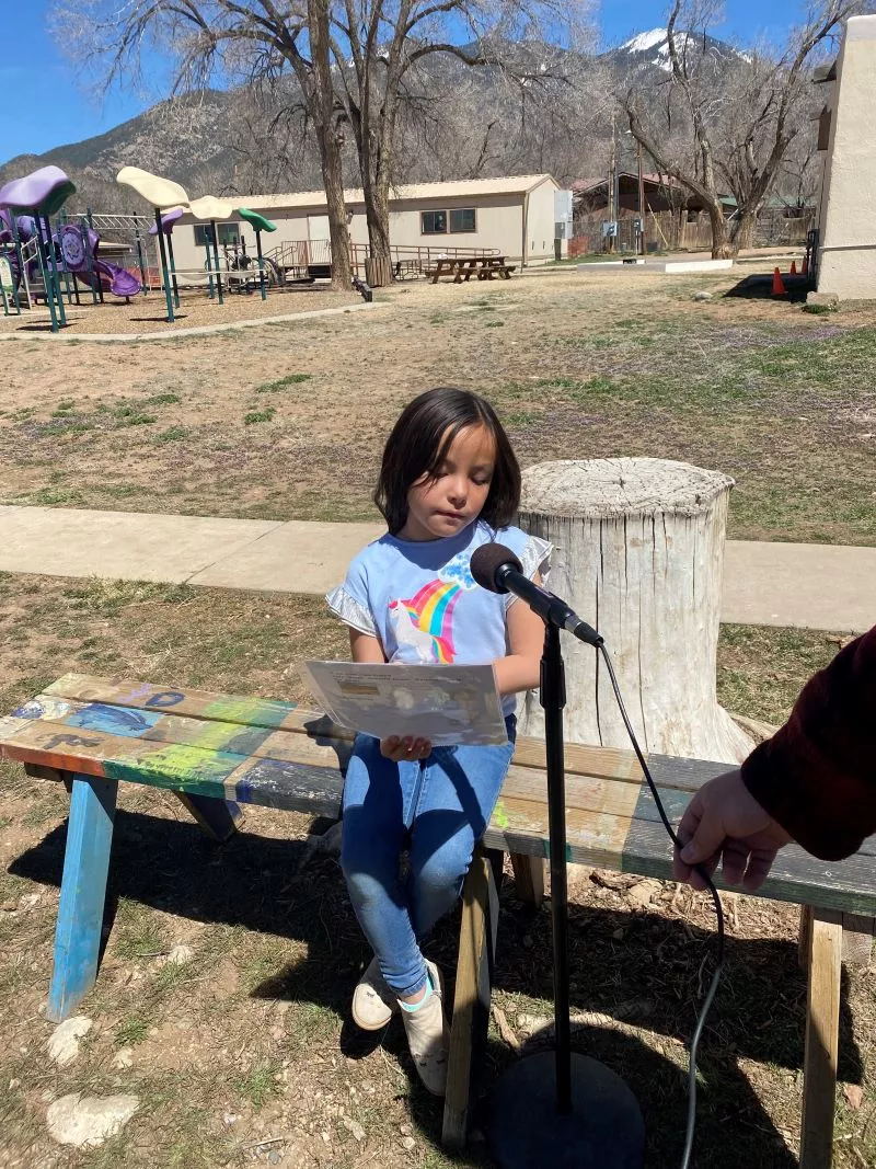 Girl Student at Taos Day School Outside Sitting on a Bench in Front of a Microphone Reading a Poem for the "I Am From Poem" Project.