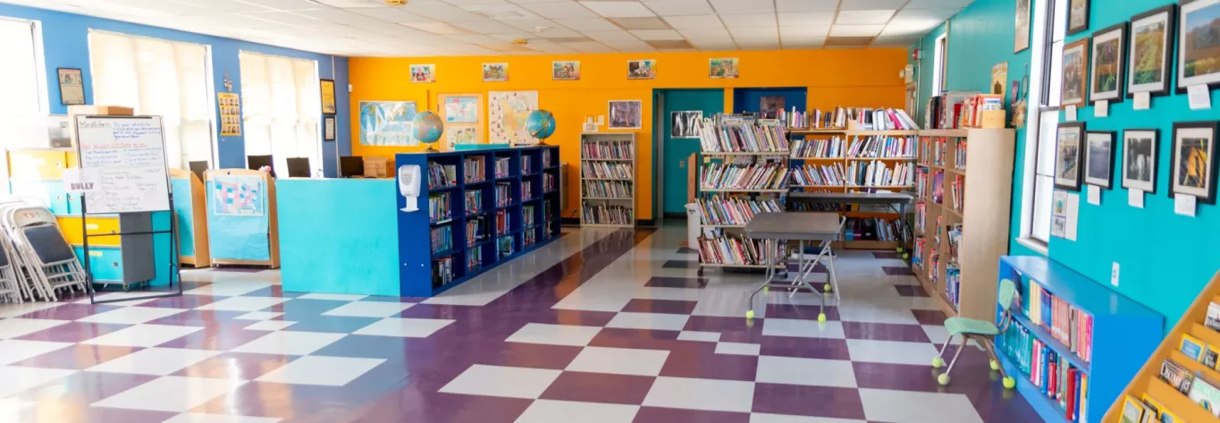 Panoramic Image of the Library with Shiney Floors, Several Shelves Full of Books, an Easel, Chairs, Desks and Wall Hangings at Taos Day School.