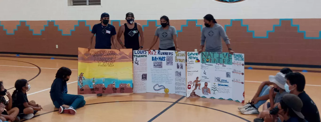 Four Taos Day School Teachers Holding Up Three Large Posterboards and Displaying Information To Students in the School Gym.