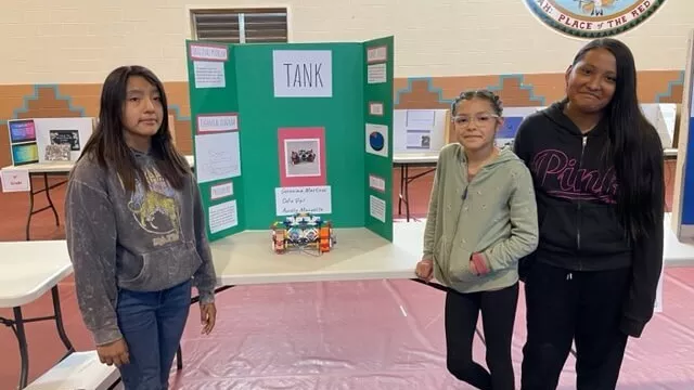 Three Female Students at Taos Day School Stand in Front of Long Table with a Trifold Project Board and Science Project Displayed Inside the Gym.