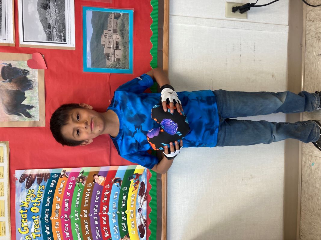 A student stands with a finished pillow sewn in class in front of a bulletin board filled with colorful notices and announcements.