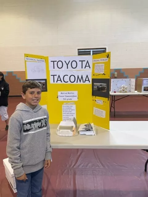A Boy Student Stands in Front of His "Toyota Tacoma" Trifold Project Board and Science Project Displayed on a Table Inside the Gym at Taos Day School.