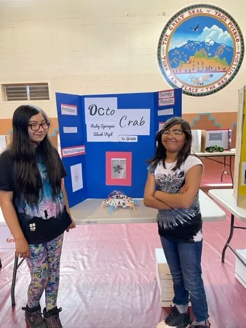 Two Girl Students Stand in Front of Their "Octo Crab" Trifold Project Board and Science Project Displayed on a Table Inside the Gym at Taos Day School.