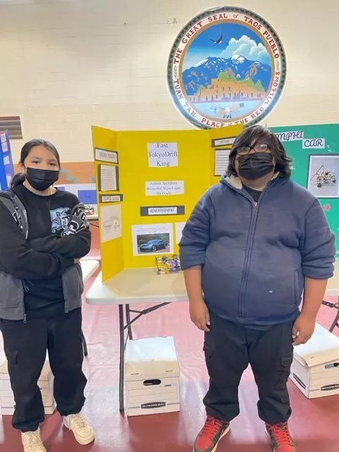 A Boy and a Girl Student Stand in Front of Their "Fast Tokyo Drift King" Trifold Project Board and Science Project Displayed on a Table Inside the Gym at Taos Day School.