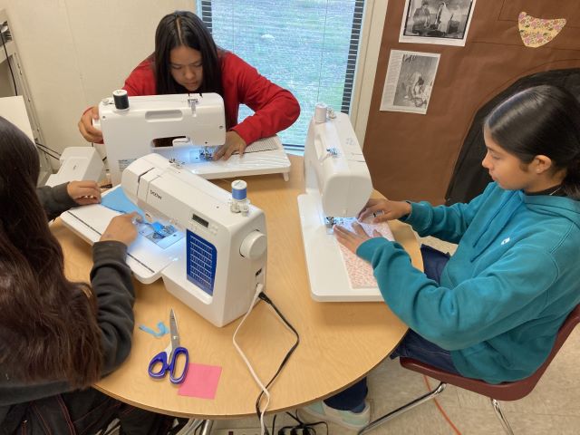 Three students sitting at a table using sewing machines to sew fabric.