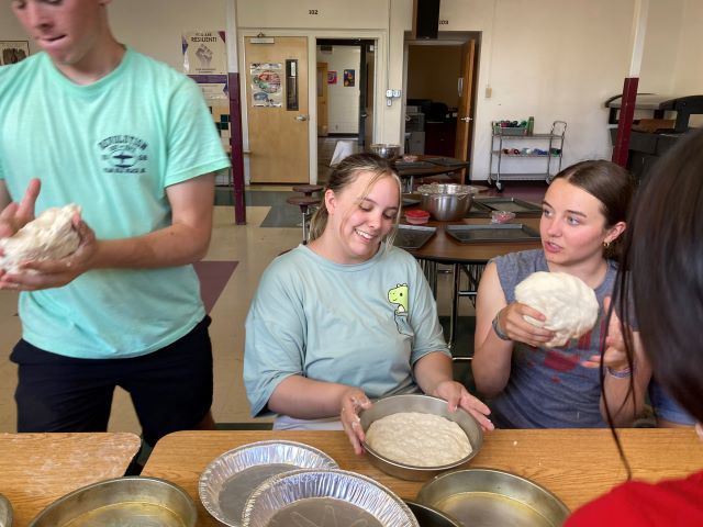 Students smiling and talking while gathered at tables preparing dough in round baking pans..