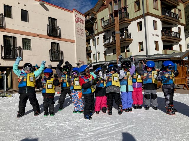 Large group of young children dressed in snowgear at ski resort covered in snow.