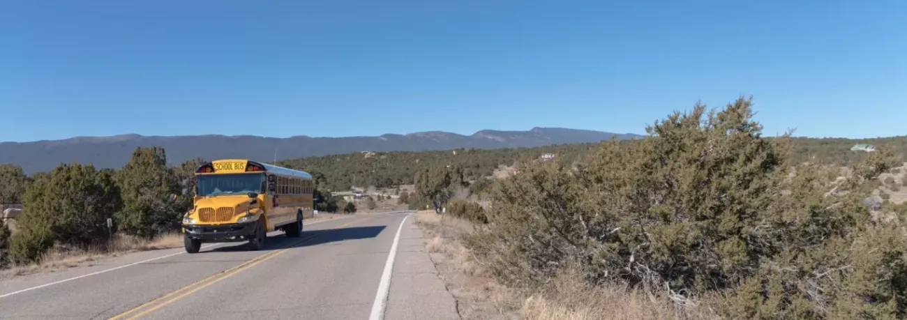 Taos Day School Yellow School Bus On Two Lane Road With Mountains and a Clear Blue Sky In The Background.