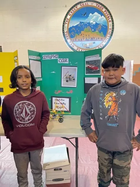 A Boy and a Girl Student Stand in Front of Their "Amphi Car" Trifold Project Board and Science Project Displayed on a Table Inside the Gym at Taos Day School.